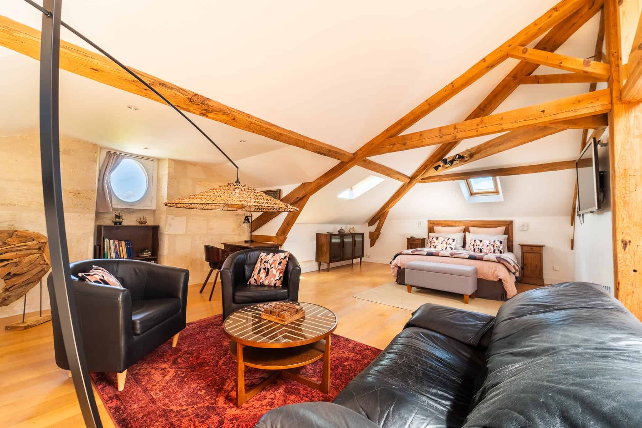 Vue intérieure de la Suite l’Estuaire avec lit queen-size, coin salon, poutres apparentes et décoration raffinée au…